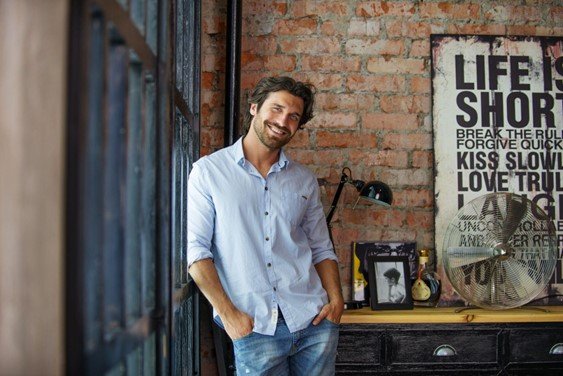 Man in a light blue shirt with rolled sleeves and jeans, leaning against a brick wall