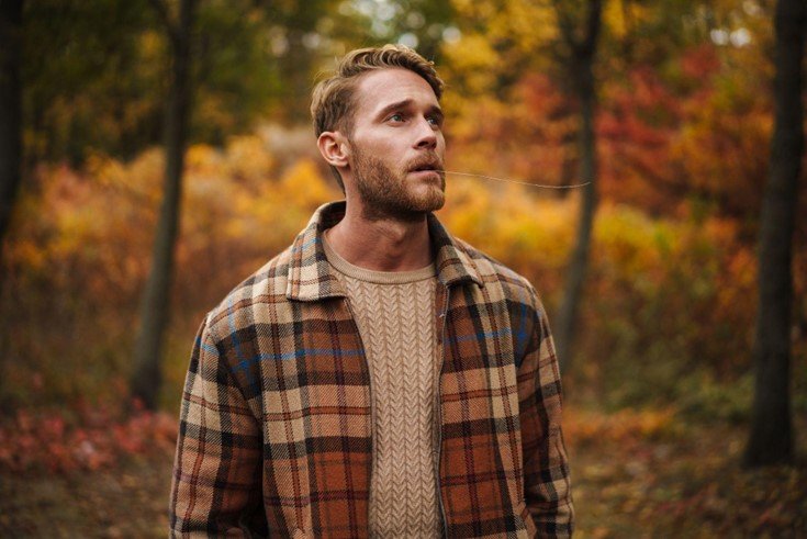 Man wearing a plaid jacket over a cable-knit sweater in a fall forest
