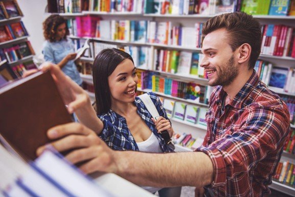 Woman turned toward a man and smiling in a bookstore reaching for the same book as they talk